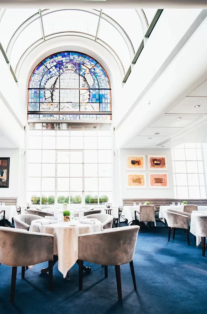 A bright, elegant restaurant interior with tall arched windows featuring stained glass at the top. The room has blue carpeting, white tablecloths, and light gray chairs. Art pieces adorn the walls, creating a sophisticated ambiance at Claude Bosi at Bibendum, London.