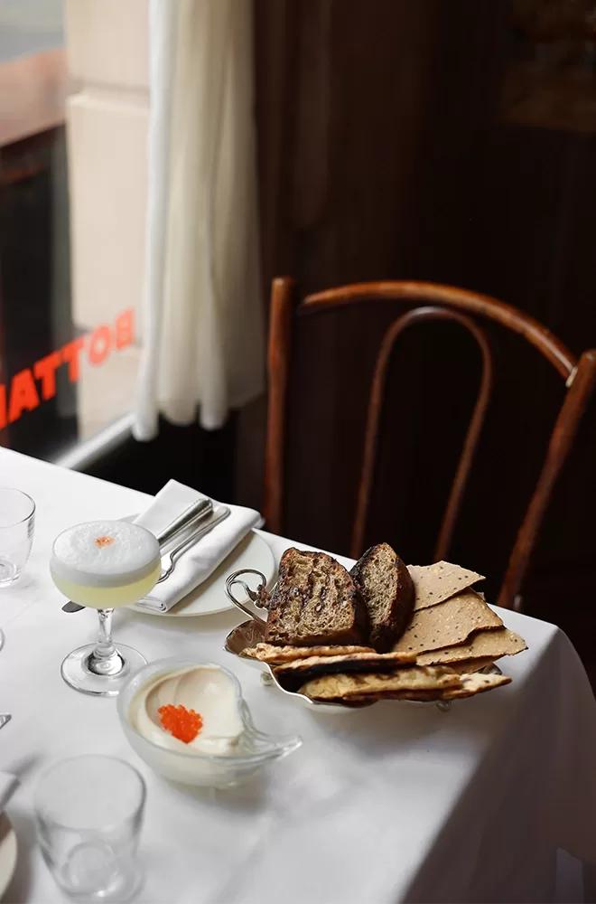 A table set with a white cloth, featuring a cocktail with foam, a bowl of creamy dip with orange garnish, and a plate of sliced bread and crackers. A wooden chair stands nearby, with an orange sign visible through the window in the background at Bottarga, London.