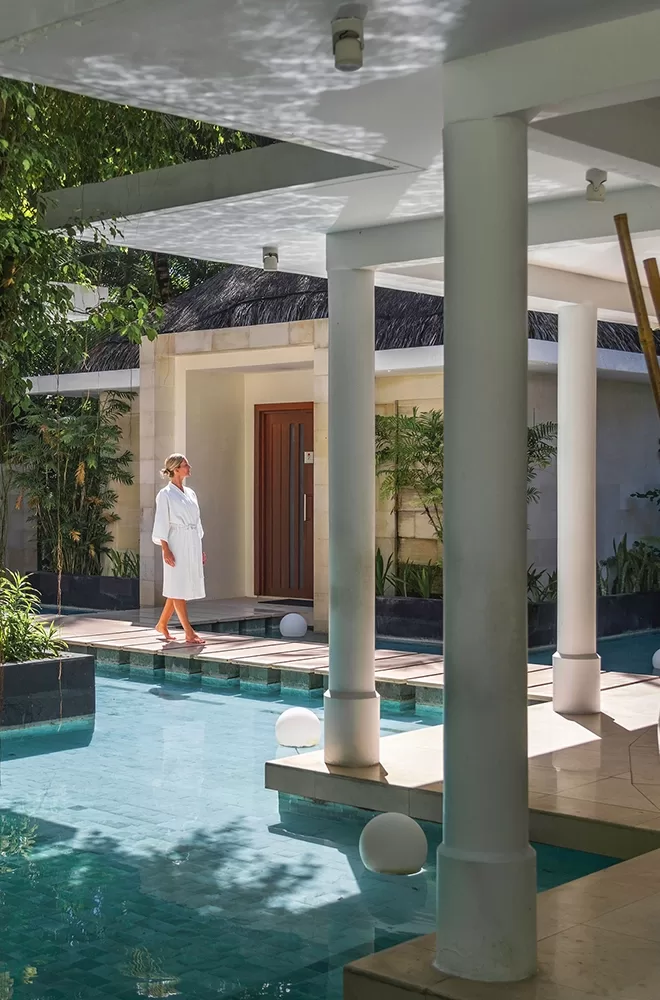A person in a white robe walks by a pool at a luxurious spa or resort, surrounded by greenery and modern architecture. The scene is serene, with a wooden door and a thatched roof building in the background at Niyama, Dhaalu Atoll, Maldives.
