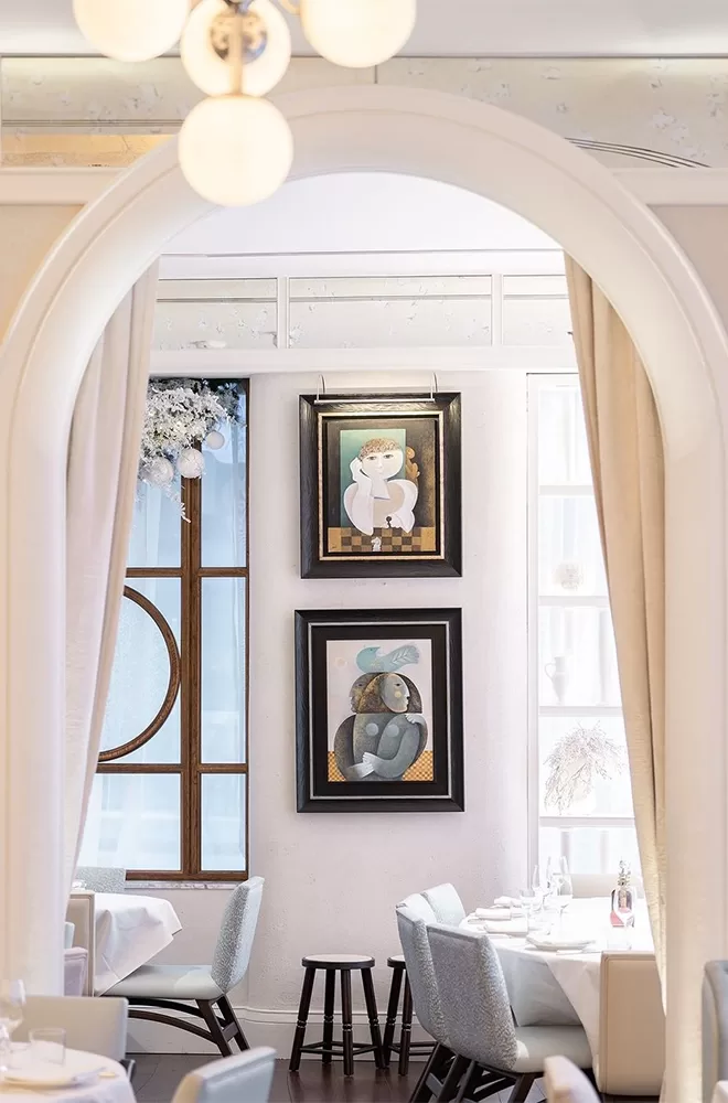 View through an arched doorway into a bright dining area with white tablecloths. Two framed paintings hang on the wall near a window. Soft lighting creates a cozy ambiance, and the room features modern gray chairs at Gaia, London.
