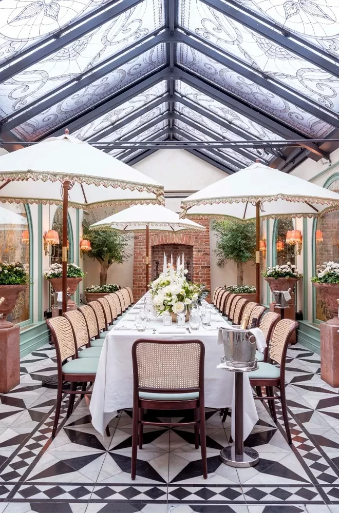 Elegant dining area with a long table set for a meal, surrounded by wicker chairs. The glass ceiling allows natural light, and large potted plants add greenery. Decorative umbrellas provide a charming touch above the table at Daphne's, London.