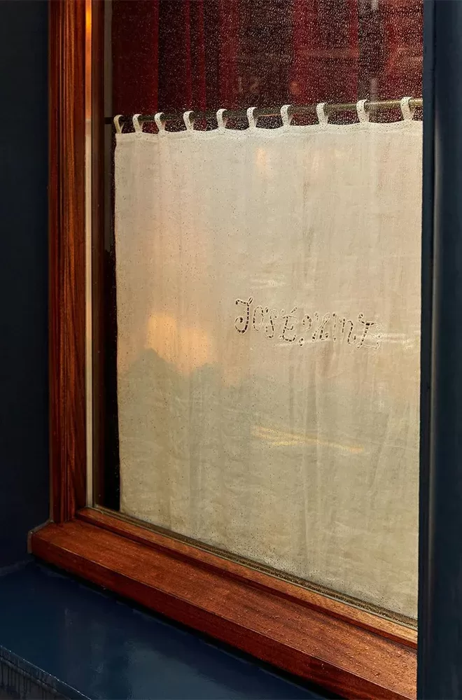 A window with a wooden frame and a light beige curtain featuring the embroidered word Joanne. The curtain is partially translucent, and the window reflects a bit of the interior at Josephine Bouchon, London.
