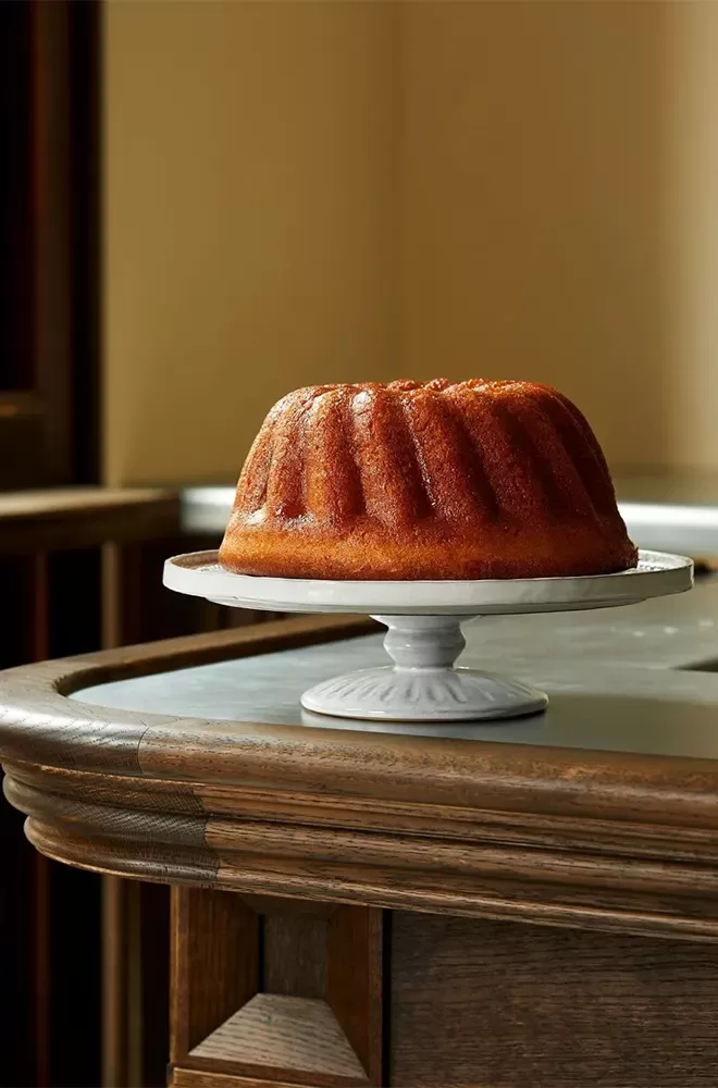 A golden-brown bundt cake sits on a white cake stand atop a wooden counter, set against a softly lit background at Josephine Bouchon, London.
