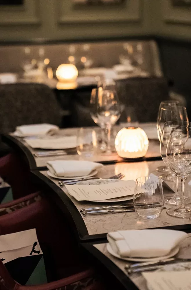 A dimly lit restaurant table set for dining, featuring wine glasses, menus, and cloth napkins. Two glowing candle holders add a warm ambiance, with more tables similarly arranged in the softly focused background at Kutir, London.