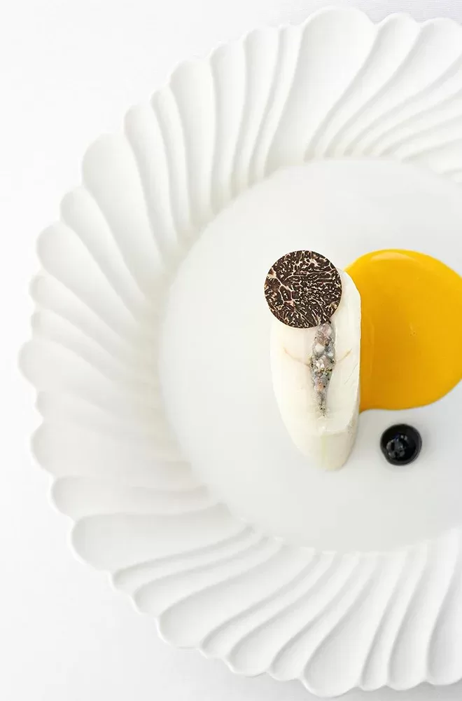 A elegantly plated dish on a white scalloped plate, featuring a cylindrical piece of fish topped with a round slice of truffle. Beside it, theres a smooth yellow sauce and a single black olive. The presentation is minimalist and artistic at Restaurant Gordon Ramsay.