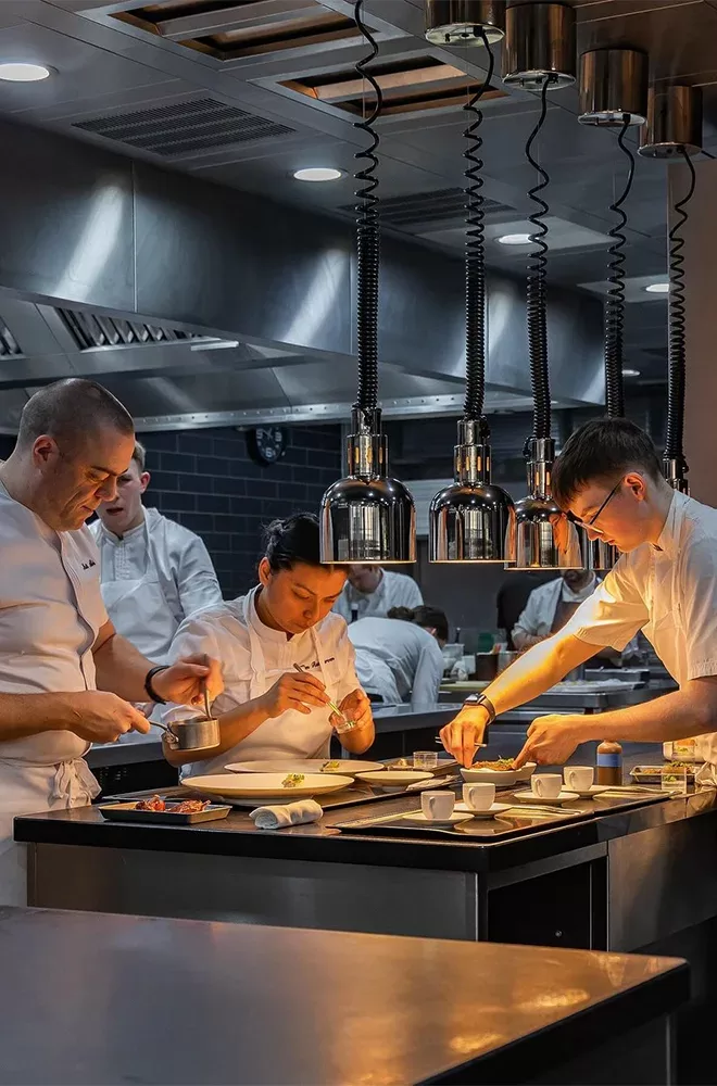 Chefs in a professional kitchen meticulously prepare dishes. The lighting is warm, highlighting the focus and precision as they work. The environment is bustling, with several chefs collaborating to create gourmet meals at Restaurant Gordon Ramsay.