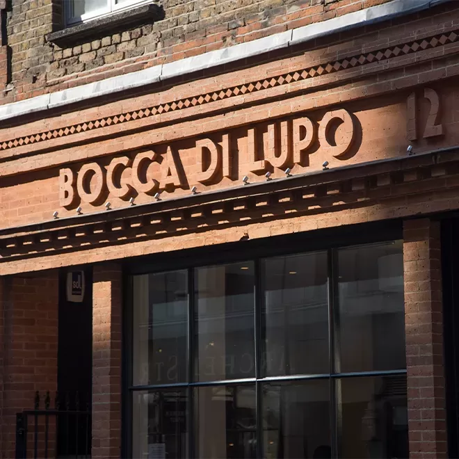 Front view of a restaurant named Bocca di Lupo, with its name in bold letters on a red brick facade. There are large glass windows and the number 12 on the building. The style is classic with an elegant touch at Bocca di Lupo, Soho, London.