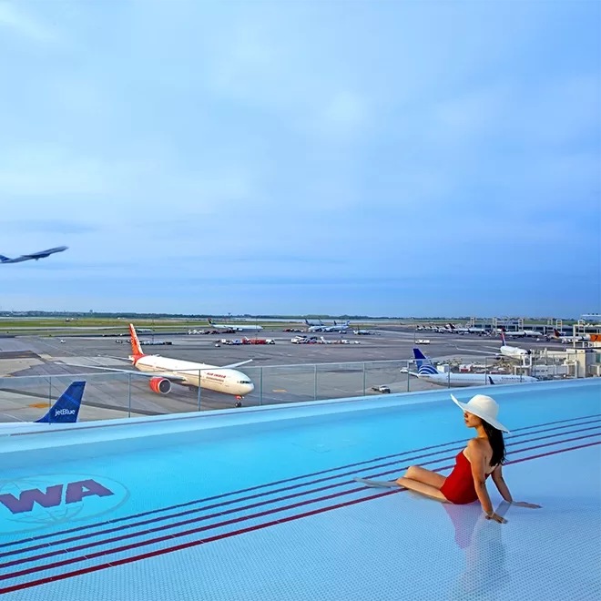 A woman in a red swimsuit and a hat dips her feet in a pool overlooking planes taking off on a runway.