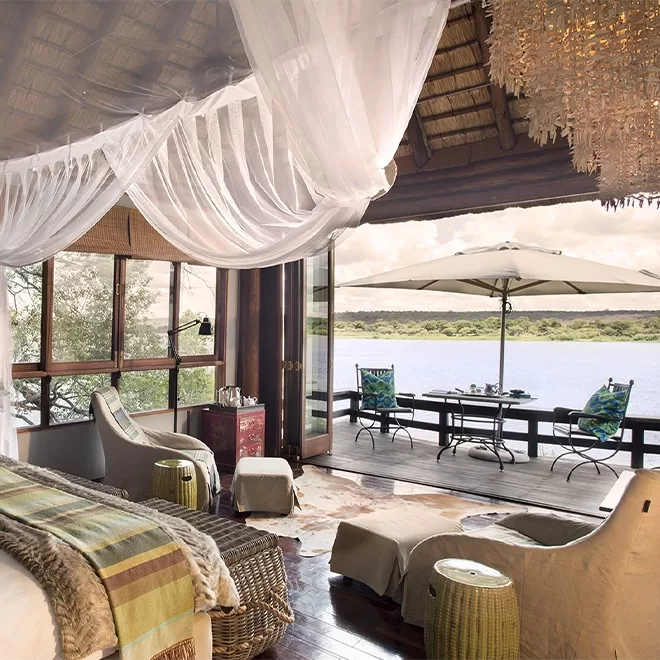 Royal Chundu Zambezi river lodge room suite interior with draped ceiling curtains, animal skin rug and chairs overlooking a balcony view of Zambia river at Royal Chundu, Zambia.