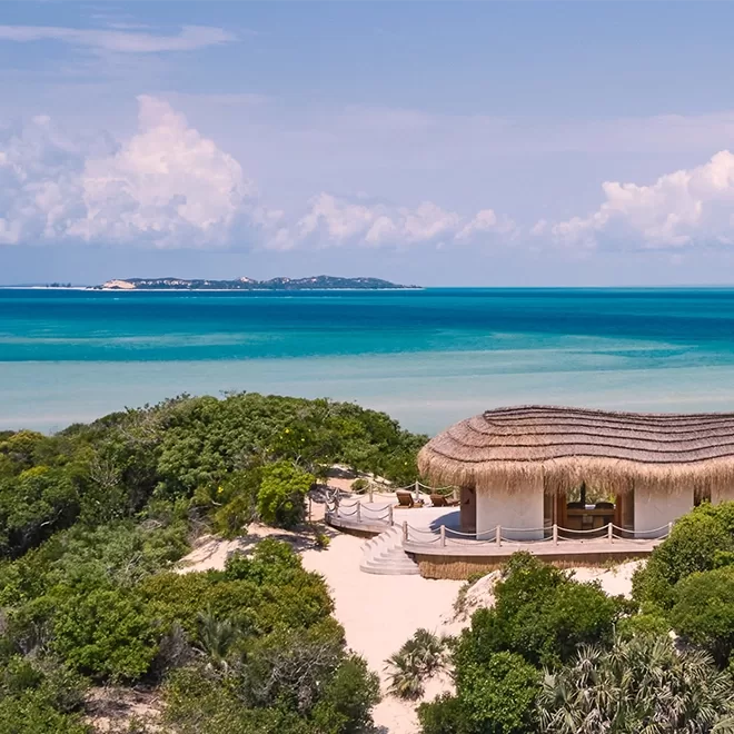 View of blue sea, white sand under straw thatched chalet and greenery at Kisawa Sanctuary, Mozambique .