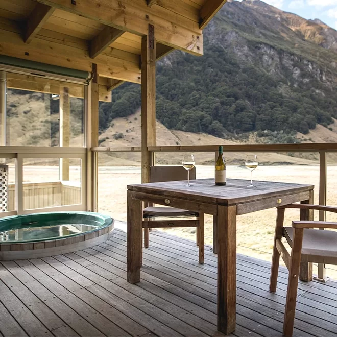 Dining table with a bottle of wine and hot tub with a view of green mountains at Minaret Station Alpine Lodge, New Zealand.
