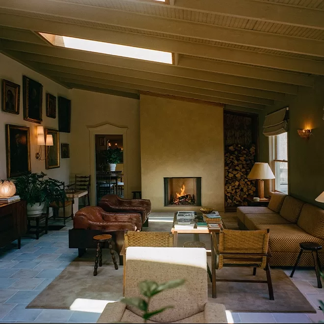 A cozy living room with a fireplace, comfortable sofas, and a leather chair. Artwork hangs on the walls, and a stack of firewood is neatly arranged. Warm lighting and plants add to the inviting atmosphere. Natural light streams in from a skylight.