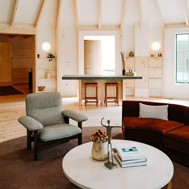 A bright, cozy room with exposed white beams, a stone wall, and an arm chair and corner sofa and a window that lets in natural light at The Rounds at Scribner’s Catskill Lodge.