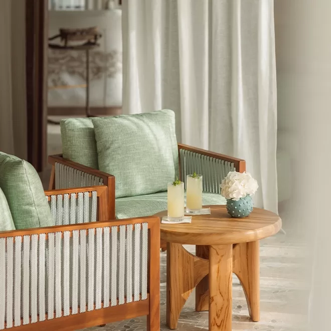 Two wooden chairs with light green cushions sit beside a small wooden round table. The table holds two tall glasses with drinks and a decorative ceramic vase with white flowers. A light curtain is in the background, creating a soft ambiance in Temuda Bay.
