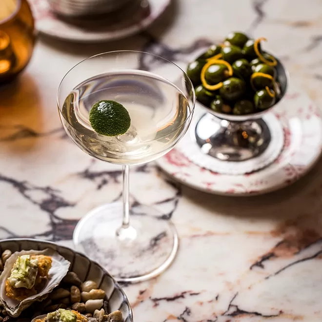 A martini glass with a clear cocktail and a lime garnish sits on a marble table. Nearby, a bowl of green olives with lemon twists and a plate of oysters with toppings can be seen. The scene has a sophisticated and elegant vibe.