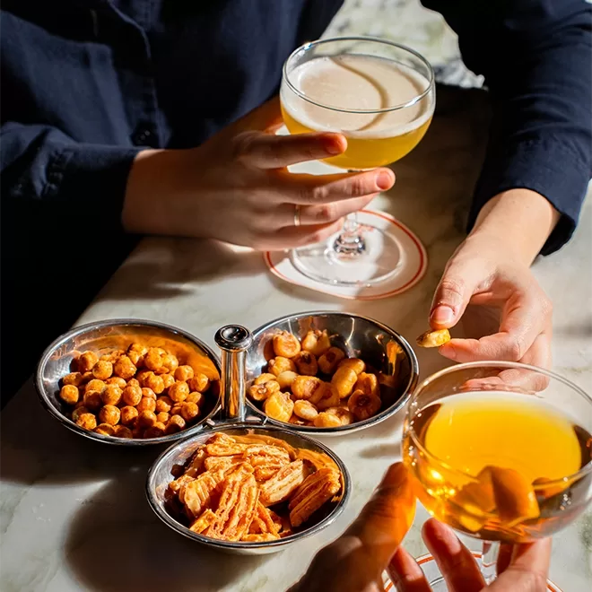 Two people holding cocktails at a table with three metal bowls containing nuts and snacks. One person reaches for a snack, while the other holds a light-colored drink. The setting appears casual and relaxed.