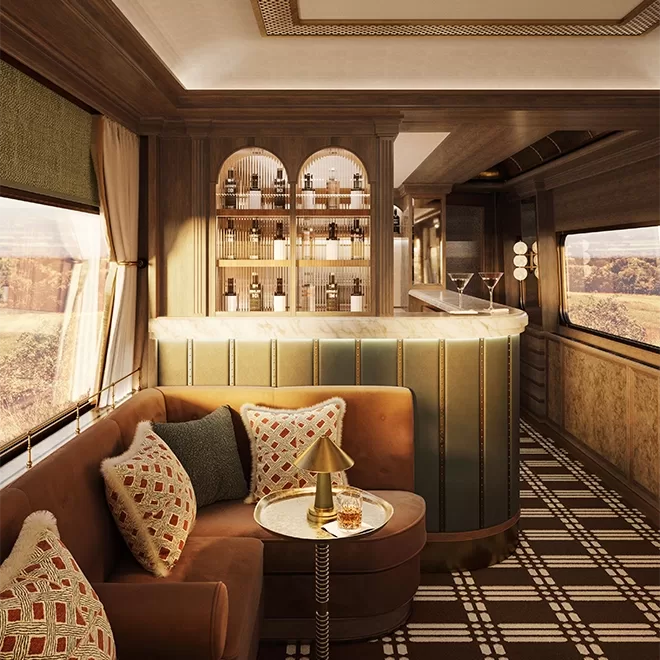 Luxurious train interior with a cozy seating area featuring a brown sofa and decorative pillows. A stylish bar with a selection of bottles and martini glasses is in the background. The decor is elegant, with a large window showcasing scenic views.