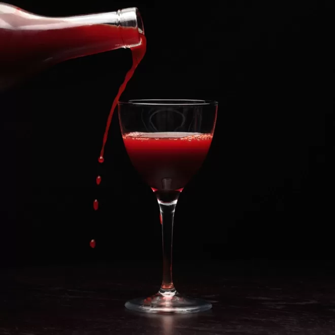 A dark red liquid is being poured from a glass bottle into an elegant wine glass against a black background, with droplets visible mid-air by Aulis, Soho.