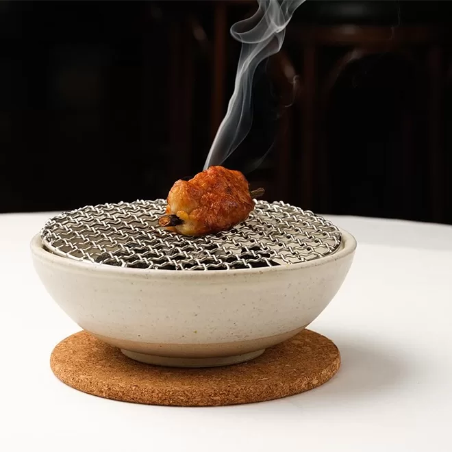 A single grilled food item on a skewer sits atop a small portable wire grill over a ceramic bowl. Smoke rises from the food, which is resting on a cork mat on a white table. The background is dark and out of focus. Served at The Clove Club, Shoreditch.