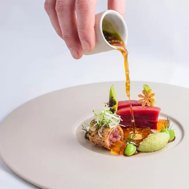 A hand pours a sauce over a gourmet dish featuring seared tuna, garnished with green herbs, small vegetables, and a puree, artistically arranged on a minimalist white plate at SO|LA Soho London, London.