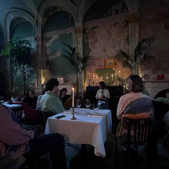 A dimly lit room with people seated around tables covered with white cloths. Candles are lit creating a warm ambiance. The walls are worn, adding an antique feel. Large plants decorate the room. A person at the front is engaged in an activity at Sessions Arts Club, Farringdon.
