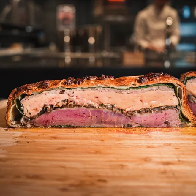 A cross-section of a beautifully crafted beef wellington-style dish, showcasing layers of tender meat, creamy filling, and a golden, crispy puff pastry crust, set on a wooden cutting board in a restaurant kitchen.