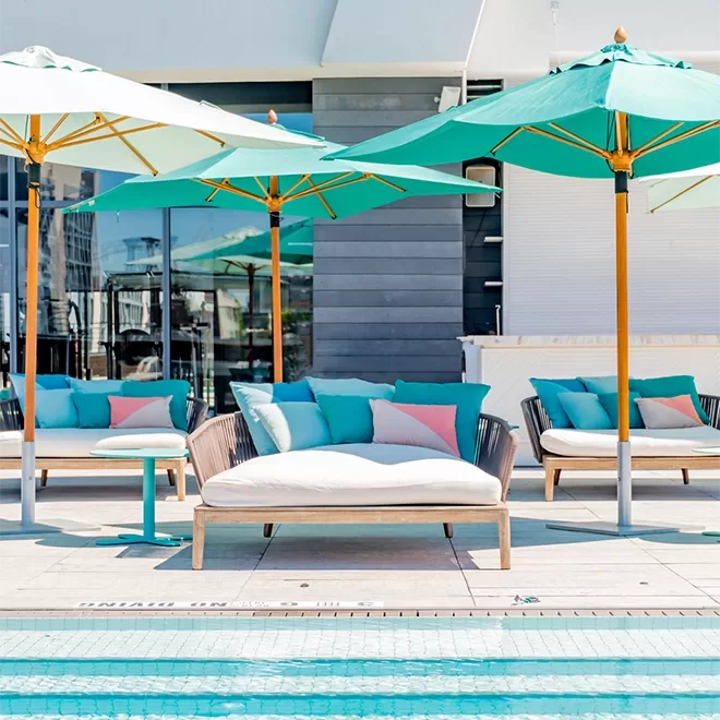Blue swimming pool edge with white, sofas facing towards the pool with baby blue and pink cushions and turquoise parasols.
