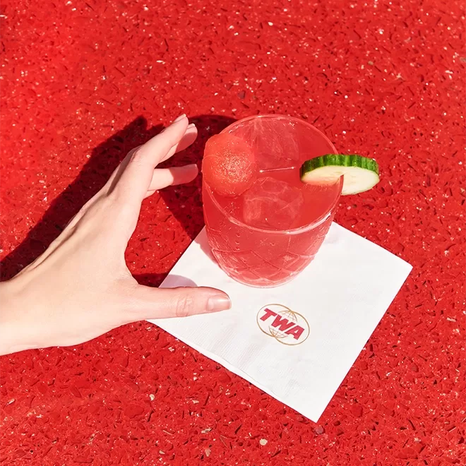 A red marble counter with a white napkin on it with a TWA logo. A refreshing cold looking glass sits on top of the napkin with a red liquid and ice with a cucumber on the side.