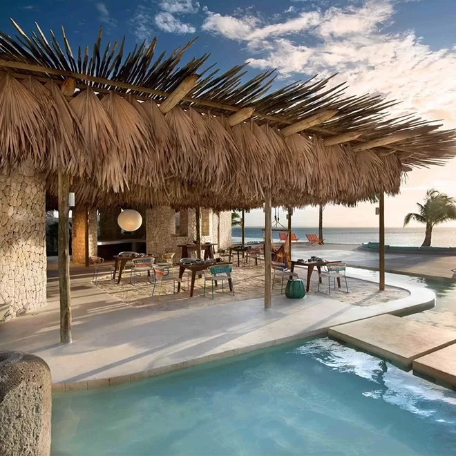A tropical beachside resort with a palm-frond-covered roof and a pool next to the ocean. The seating area under the thatched roof is furnished with colorful chairs at Miavana by Time + Tide, Madagascar.