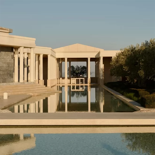 A luxurious, minimalist pavilion with white columns and a long reflective pool in front. The structure has classical elements and is surrounded by greenery, at Amanzoe, Greece.