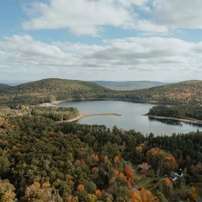 Aerial view of a serene lake surrounded by dense forests in autumn colors. The landscape features rolling hills and a partly cloudy sky, capturing the essence of a tranquil, natural setting.