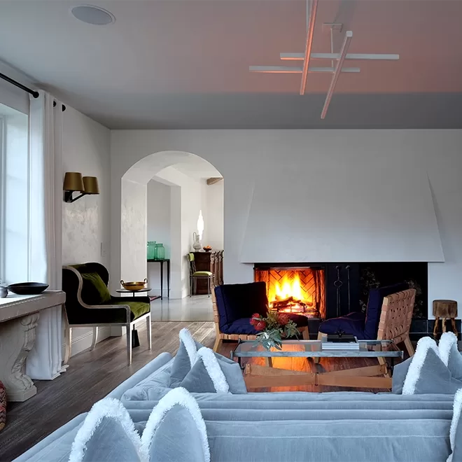 A cosy living room at The Villa at Saugerties boasts modern decor with a lit fireplace, velvet chairs, and a glass coffee table. Light gray walls, wooden flooring, and a blend of contemporary and classic furniture are enhanced by soft lighting that creates a warm ambiance.