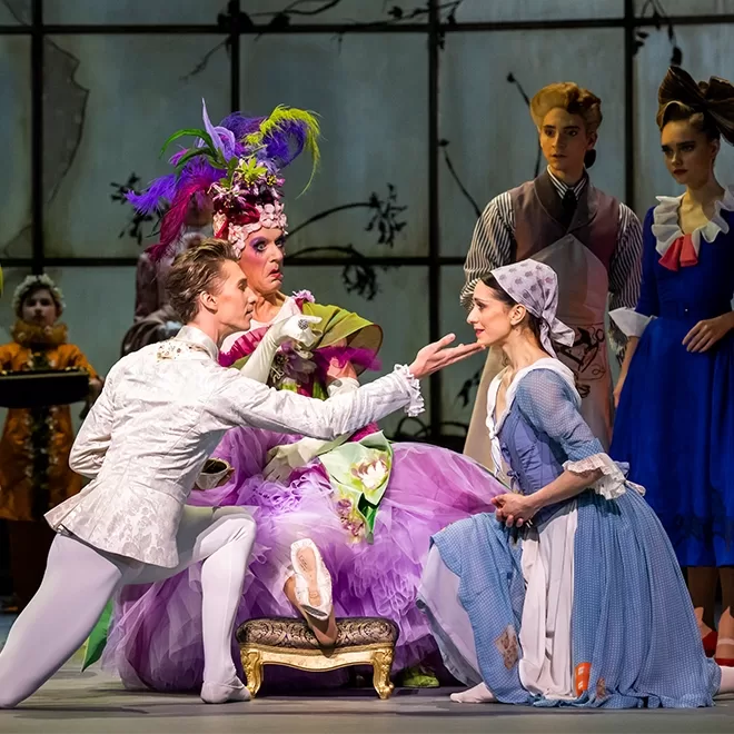 Ballet dancers perform in an ornate theatrical scene. A dancer in a flamboyant purple costume with a towering headdress interacts with a dancer dressed in a blue peasant outfit, while others in elaborate attire watch at Cinderella.