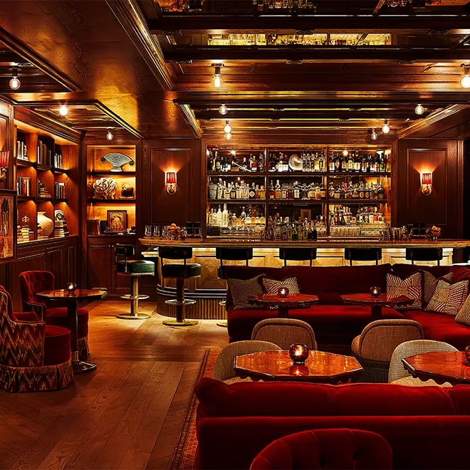 A warmly lit, elegantly designed bar interior featuring plush red seating, wooden floors, and a well-stocked bar with high stools. Soft ambient lighting and decorative shelves create a cozy and inviting atmosphere.
