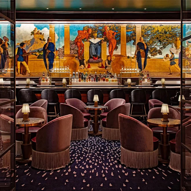 A luxurious bar interior featuring plush pink velvet chairs, round tables with lamps, and a mural depicting figures and nature scenes. The countertop displays various bottles, creating an elegant and opulent atmosphere.
