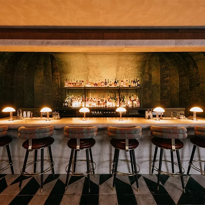 A dimly lit bar interior with a row of cushioned bar stools in front of a marble counter. Warm, glowing lamps line the bar, and shelves behind are stocked with various bottles. The floor has a black and white geometric pattern.