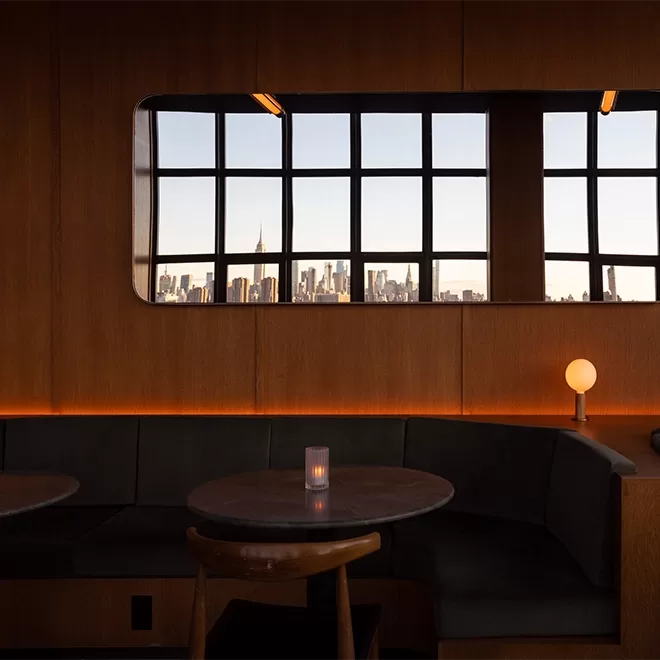 A cozy, dimly lit lounge with a curved booth and round table, featuring a candle. A large wall mirror reflects a city skyline through grid windows, enhancing the warm wooden decor.