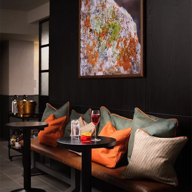 This cosy lounge at Aulis, Soho, features a cushioned bench with colorful pillows, a small table with drinks, and a vibrant wall art piece. Soft lighting creates a warm ambiance, with a drinks cart visible in the background.