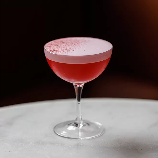 A cocktail served at The Clove Club, Shoreditch, in a coupe glass sits on a white marble surface. The drink is red with a frothy, pink layer on top, sprinkled with red dust. The background is dark, highlighting the vibrant color of the cocktail.