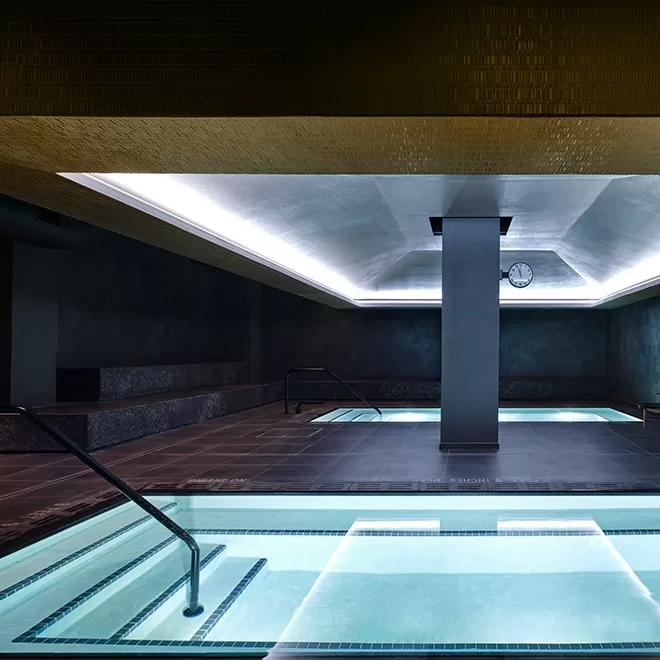 A dimly lit indoor swimming pool with a modern design featuring a small connected hot tub. The space has sleek, dark walls and a large rectangular light fixture on the ceiling. Several stair railings lead into the pool. A clock is visible on a pillar.