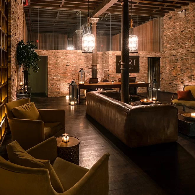 Cozy, dimly lit lounge with brick walls, featuring brown leather couches, yellow armchairs, and candlelit tables. Two large chandeliers hang from the wooden ceiling. Theres a potted plant and wooden furniture, creating a warm, inviting atmosphere.