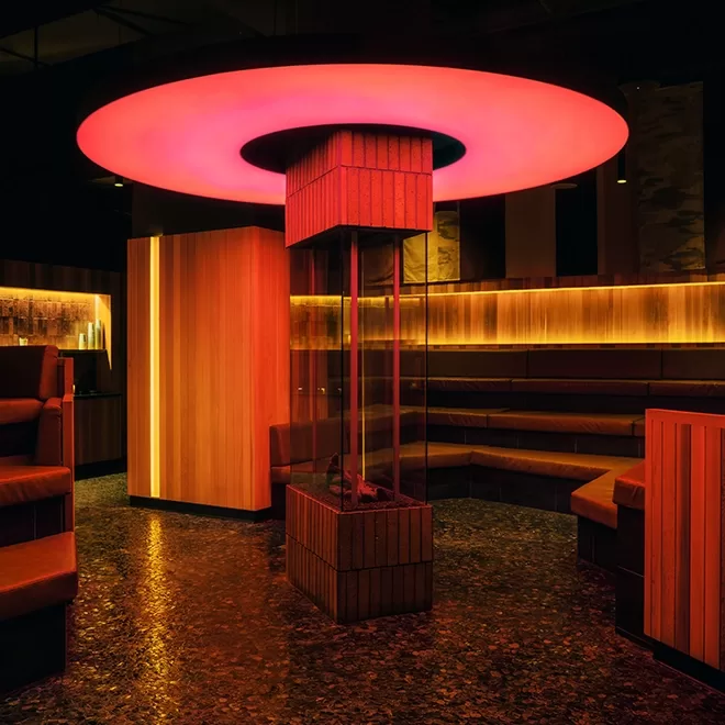 A modern lounge area with a central column featuring a red glowing circular light fixture above. The room has wooden paneling and seating arranged around the central structure, creating a warm and inviting atmosphere.