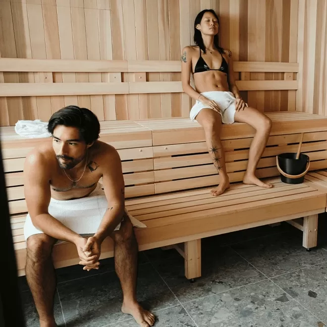 A man and woman are sitting in a wooden sauna. The man, wearing a towel around his waist, sits with his hands clasped. The woman, wearing swimwear, sits comfortably with a towel on the bench and a bucket nearby.