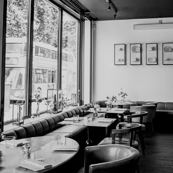 Black and white image of a modern cafe with large windows. A bus passes by outside. Inside, round tables with chairs are set with glassware and flowers. Framed art decorates the white wall at Angelina, Dalston.