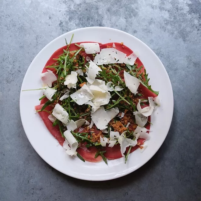 A white plate with thinly sliced beef carpaccio topped with arugula, shaved Parmesan cheese, and crispy breadcrumbs, set on a gray textured surface at Jolene, Newington Green.