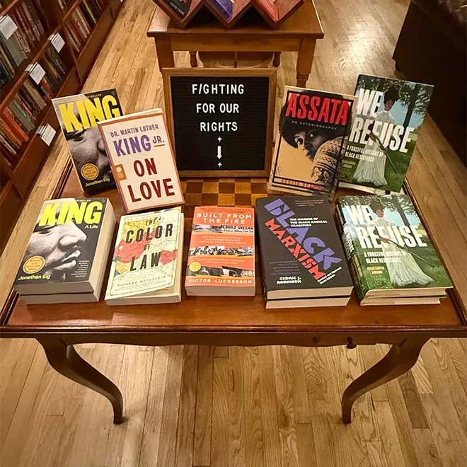 A wooden table displays various books on social justice and activism at Liz's Book Bar, Brooklyn, NYC. Behind them, a letter board reads Fighting for our rights with an arrow pointing down. The table is set in a bookstore with a wooden floor.