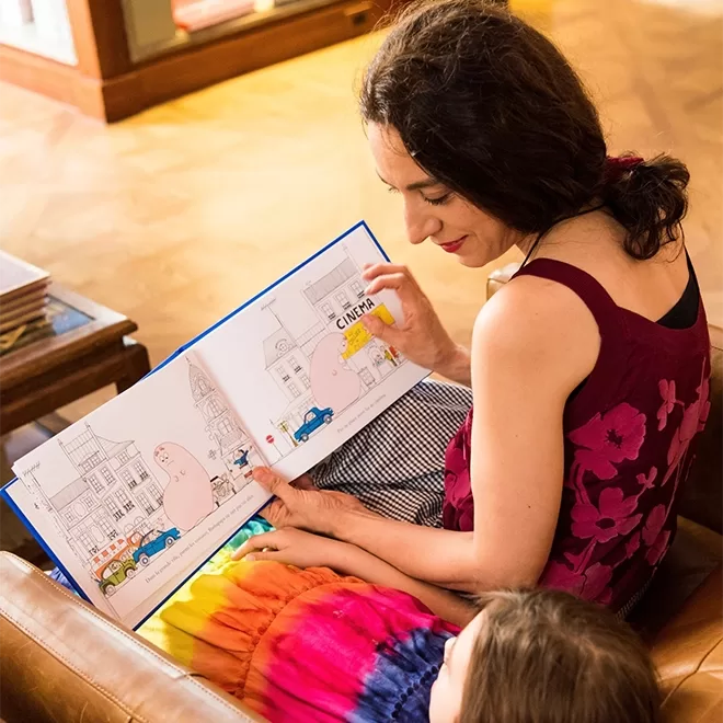 Woman in a floral top reads a colorful childrens book with a child. They sit on a brown leather couch inside Albertine bookstore, Upper East Side, in NYC. The book shows a city street with cars and a cinema. Bright room with wooden flooring and a bookshelf in the background.