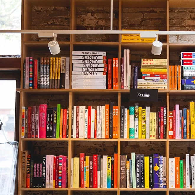 A wooden bookshelf inside Books Are Magic in NYC filled, with a variety of colorful books. Each shelf displays books of different genres and colors, creating a vibrant and visually appealing pattern. Two small spotlights are attached above, illuminating the collection.