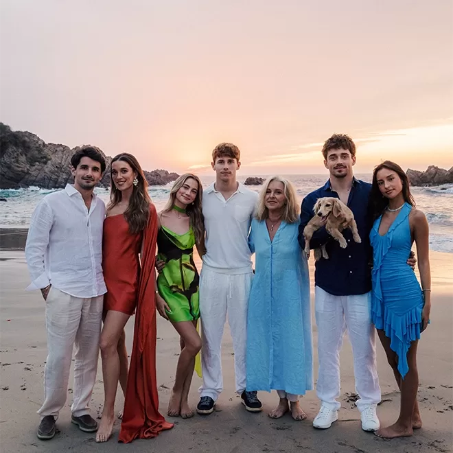 Seven people stand on a beach at sunset, smiling and posing together among them, Charles Leclerc and his family. One person holds a small dog. Everyone is dressed in summer outfits, with a mix of dresses and casual shirts and trousers. The ocean and a rocky coastline are visible in the background.