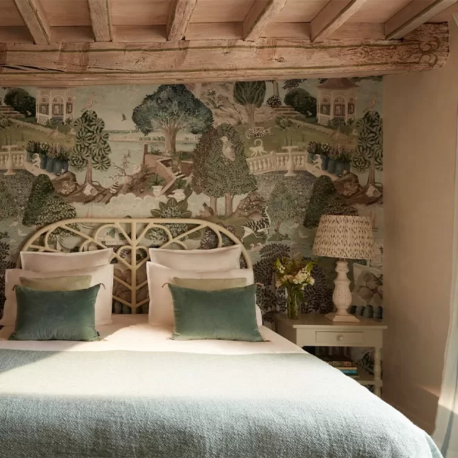 A cosy bedroom with a large bed featuring green and white pillows and a teal blanket. The wall is covered with a scenic wallpaper depicting trees and houses. A rustic wooden beam ceiling is above. A small table with a lamp and flowers is by the bed in France.